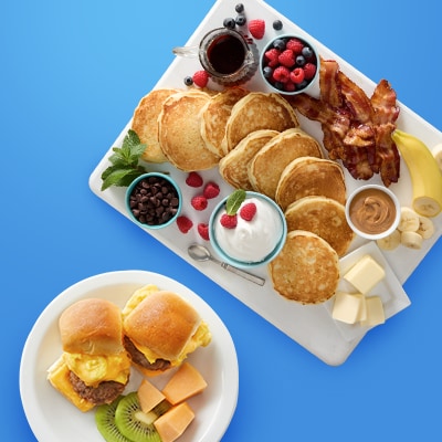 A breakfast spread on a blue background featuring pancakes, bacon, berries, chocolate chips, whipped cream, peanut butter, banana slices, and butter cubes on a platter. Nearby is a plate with breakfast sliders, kiwi slices, and cheese cubes.