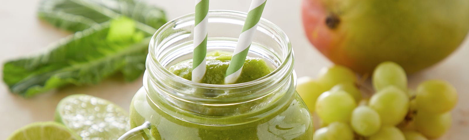 A green, St. Patrick's Day inspired smoothie in a mason jar with two straws surrounded by collard greens, grapes and a lime