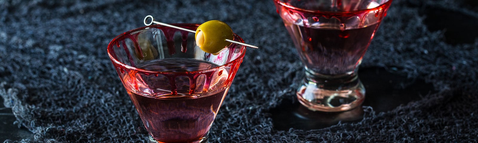 Two cocktail glasses with red liquid dripping down their sides sit on a dark blue cloth
