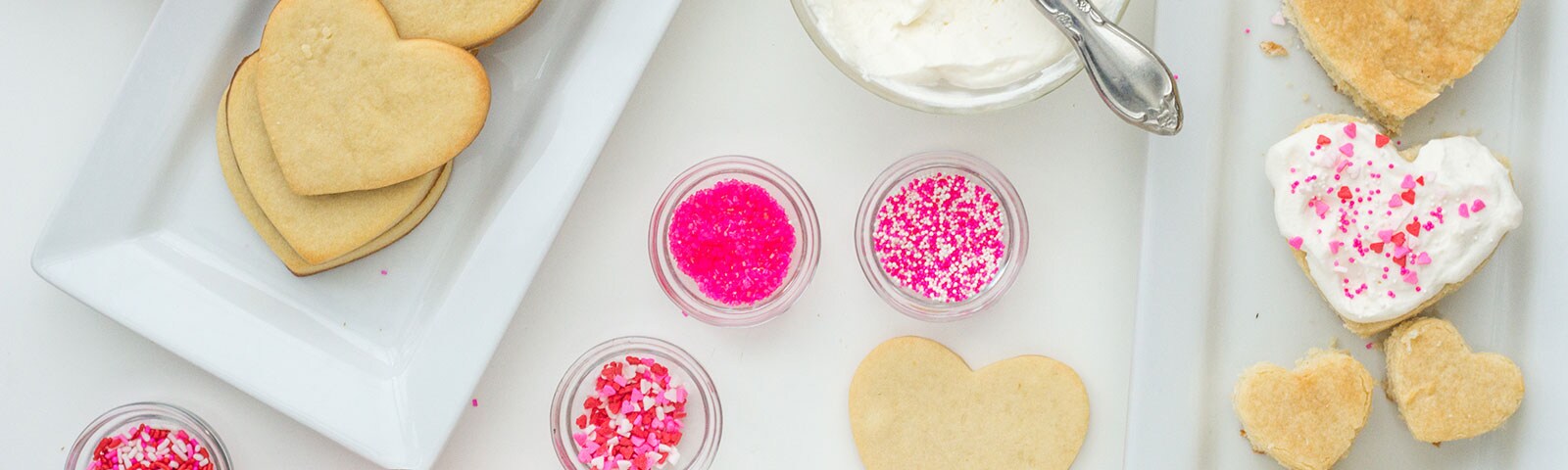Heart-shaped sugar cookies on a tray next to pink sprinkles and white frosting in separate bowls