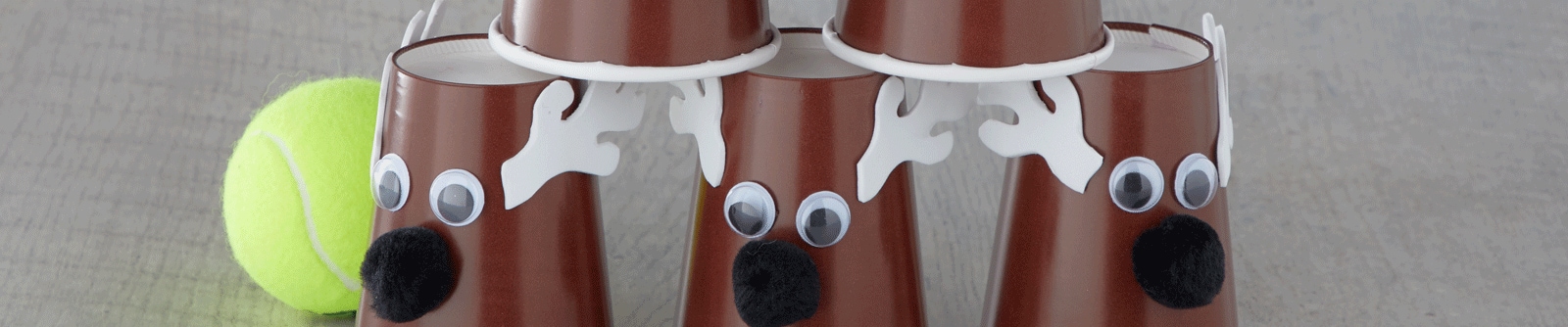 A stack of DIY reindeer using brown plastic cups with googly eyes, felt antlers and pompoms next to a tennis ball