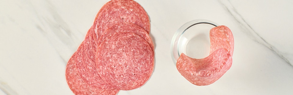 Salami slices layered over wine glass rim, on a white marble surface.
