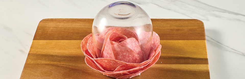 Wine glass flipped upside down with salami slices layered underneath in a rose shape. Wine glass is on a wooden cutting board, on a white marble surface.